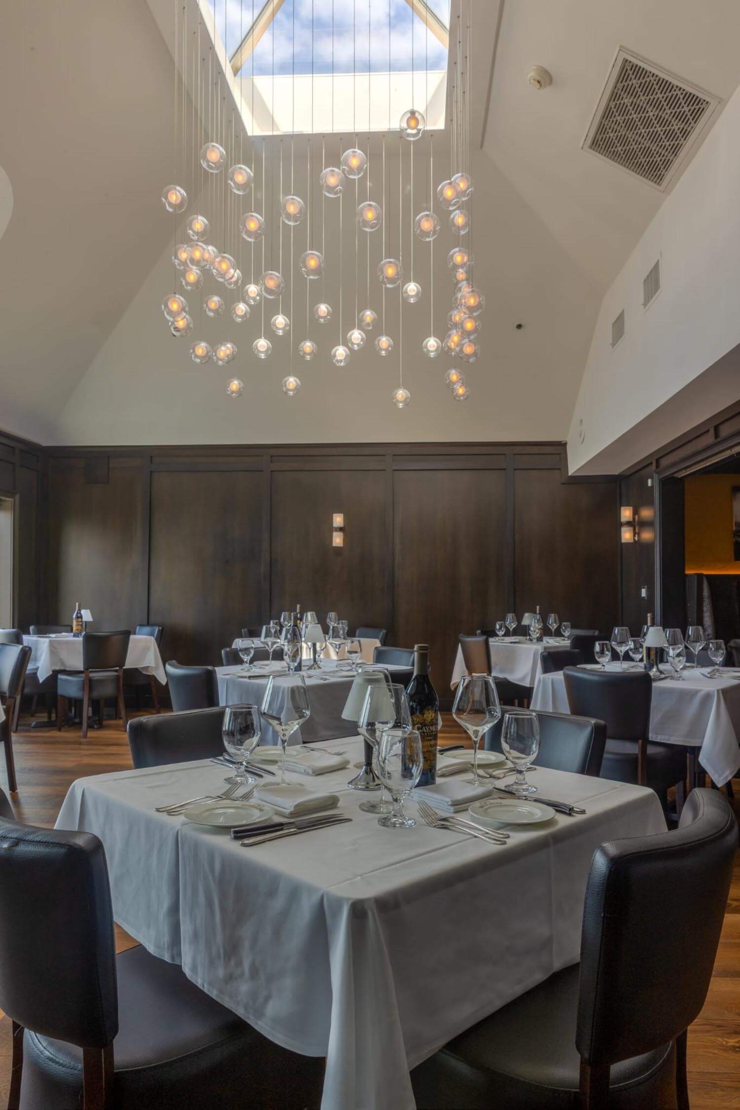 Elegant restaurant interior with white tablecloths, wine bottles, glassware, and modern hanging lights under a skylight.